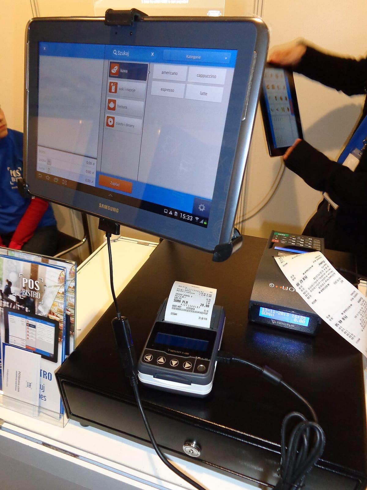 A tablet mounted as a point-of-sale terminal, with a small receipt printer and a cash drawer on the counter below it