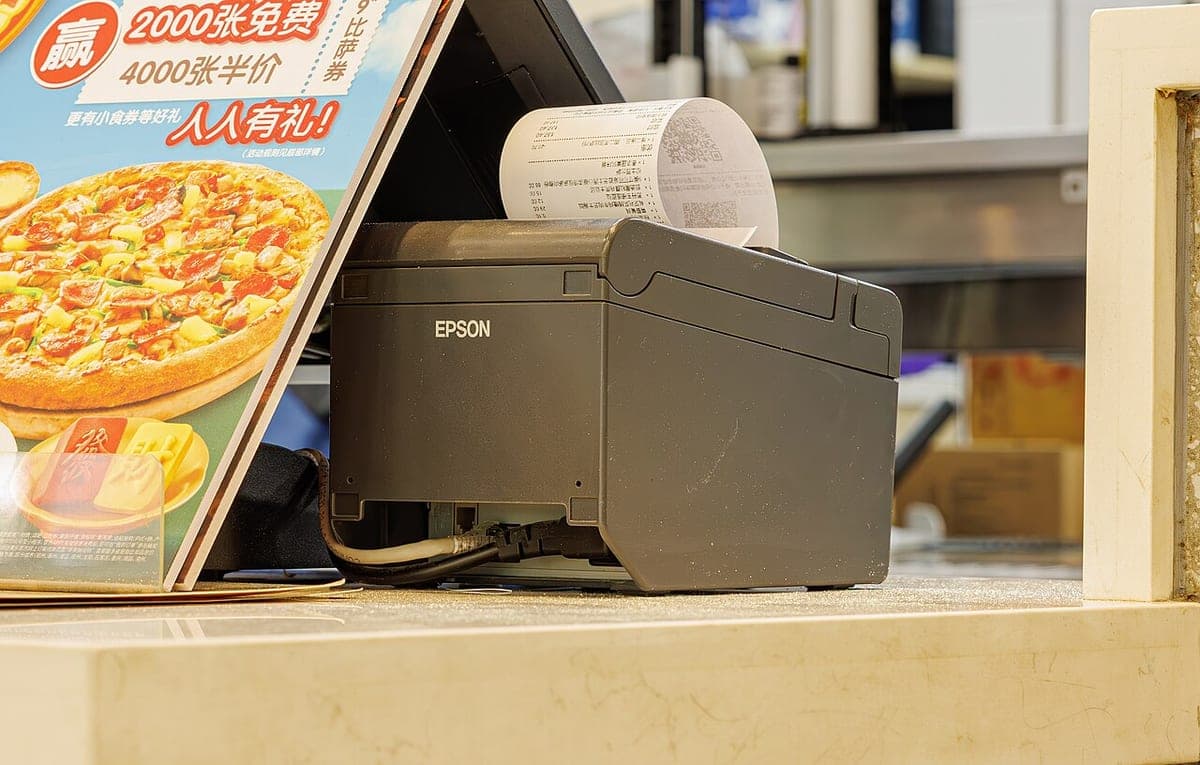 Printer struk Epson difoto dari depan, memperlihatkan penutup kertas dan tombol status
