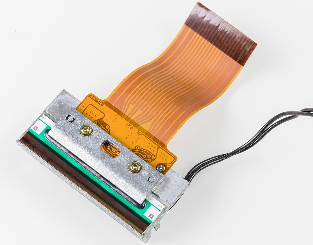 Close-up of a thermal print head module showing the heating element strip and its orange flexible ribbon cable