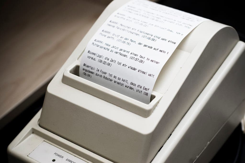 A small thermal receipt printer on a desk with a printed receipt curling out of its paper slot